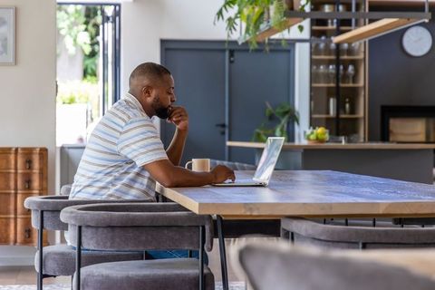 African man working from home at modern open-plan kitchen table with laptop and coffee
