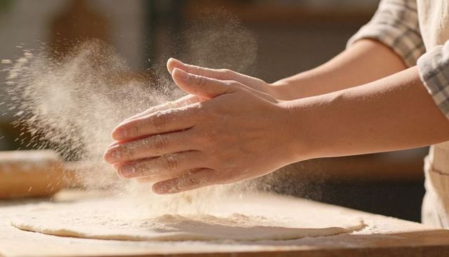 Baker hands dusting flour over dough on wooden board, artisan baking, rolling pin nearby