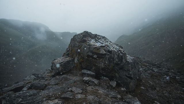 Misty mountain boulder sitting on rugged pass, snow flurries falling, lichen-covered rock