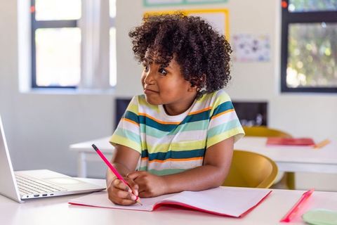 Curious african american schoolboy studying with laptop and notebook in bright classroom