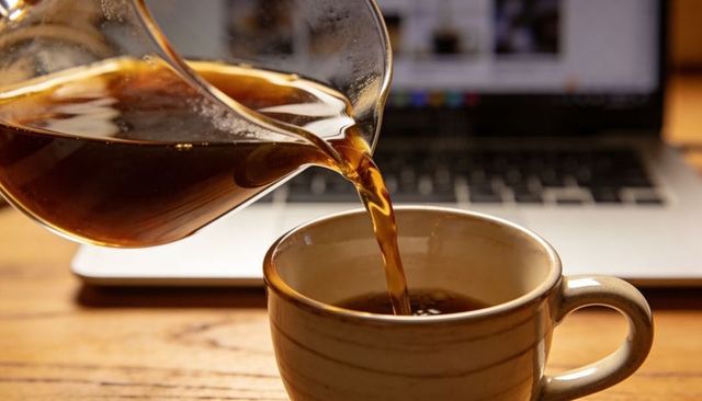 Pouring fresh coffee from glass carafe into beige ceramic mug on wooden desk with laptop for work