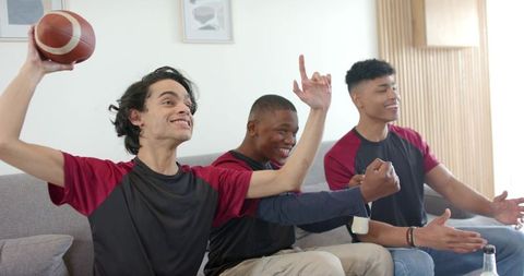 Diverse friends cheering while throwing football on couch during game night