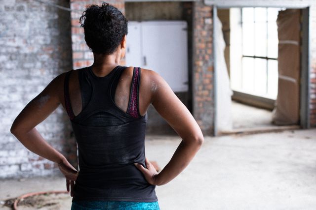 This image depicts an African American woman in sports clothes taking a break in an empty urban building. Ideal for use in fitness and health-related content, motivational materials, urban lifestyle promotions, and advertisements focusing on strength and determination.