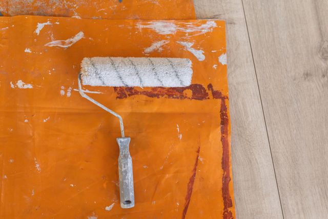 High angle view of paint roller with white paint on orange cloth on wooden floor. DIY painting decorating renovating project.