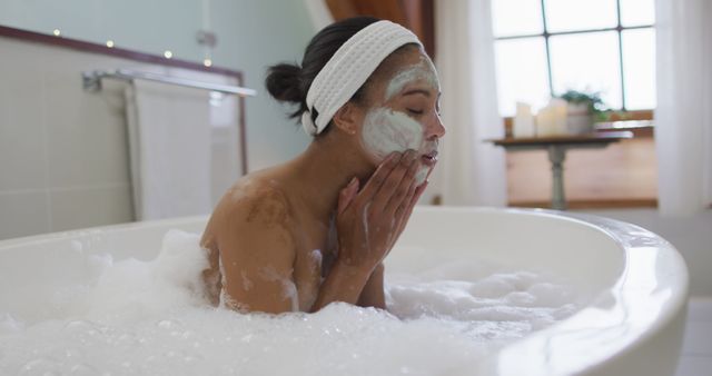 Biracial woman sitting in bathtub taking a bath, cleaning her face. domestic life, spending quality free time relaxing at home.