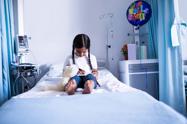 Young girl sitting on hospital bed using digital tablet, surrounded by medical equipment and a 'Get Well Soon' balloon. Ideal for use in healthcare, pediatric care, technology in medicine, and recovery-related content.