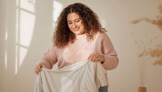 Curly-haired woman folding white shirt smiling in cozy sunlit home interior relaxed morning