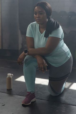 African american woman kneeling on gym floor wearing teal activewear and smartwatch