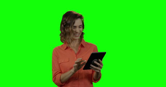 Smiling woman in orange shirt using tablet with green screen background. Ideal for presentations, marketing, technology promotions, website headers, and social media content showcasing modern digital lifestyles and device interactions.