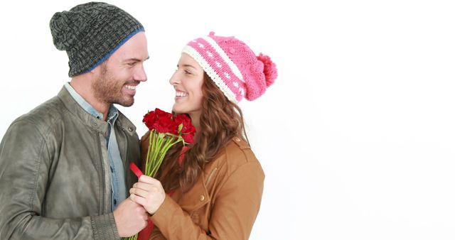 A Caucasian man and woman share a romantic moment, with the man presenting the woman with a bouquet of red roses, with copy space. Both are dressed in warm winter clothing and beanies, suggesting a cozy, affectionate atmosphere.