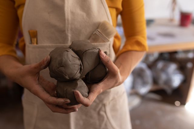 Caucasian female potter holding a lump of clay in a pottery studio. She is wearing an apron and standing at a work table with shelves in the background. Ideal for use in articles or advertisements about pottery, art, creativity, handmade crafts, and ceramic workshops.