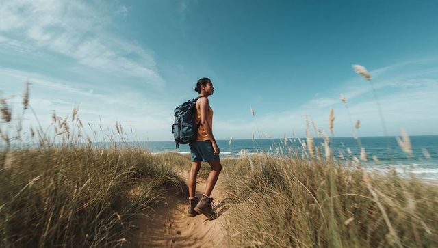 Solo hiker standing on coastal dune path facing ocean with backpack and hiking boots