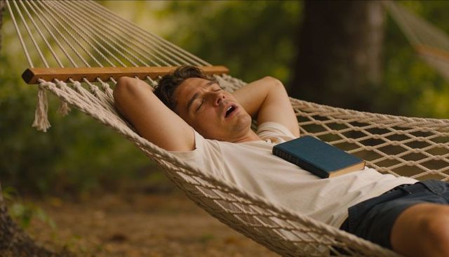 Young man napping in rope hammock with book sunlit park tranquil outdoor relaxation