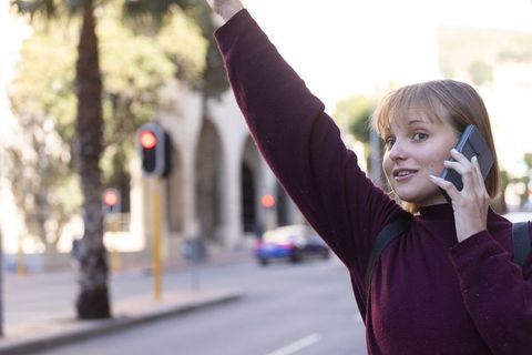 Young woman hailing taxi while talking on smartphone on sunny urban street