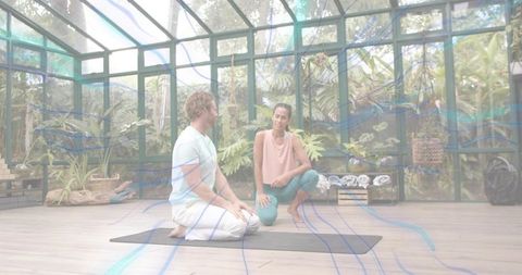 Yoga instructor guiding kneeling student in glass greenhouse studio for mindful balance
