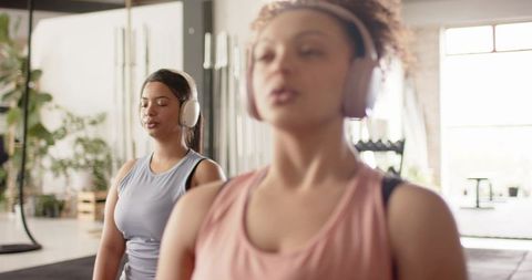 Two women stretching with wireless headphones in airy gym near dumbbells and plants