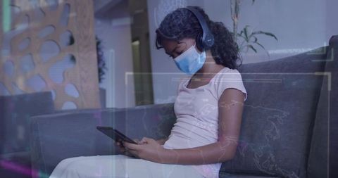 Young student wearing mask and headphones using tablet on couch for remote learning