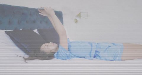 Woman lying on bed using smartphone while wearing blue pajamas in minimalist bedroom
