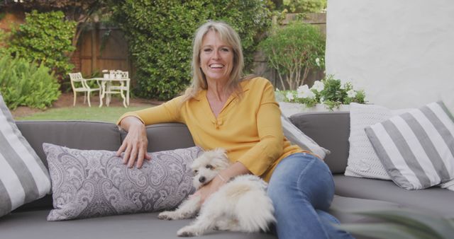 A middle-aged woman is relaxing on an outdoor couch in a backyard garden while lovingly holding her small dog. She is casually dressed in a yellow shirt and jeans, emanating a sense of contentment. The garden is lush and green, and there's patio furniture in the background. This image can be used for promoting outdoor furniture, pet companionship, relaxation, and lifestyle-related content.