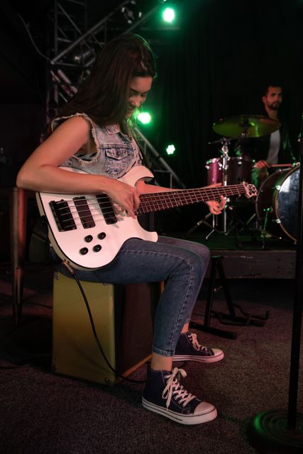 Caucasian woman sitting and playing electric guitar with male drummer and band equipment on stage at a music venue during concert in the background. Entertainment fun music.