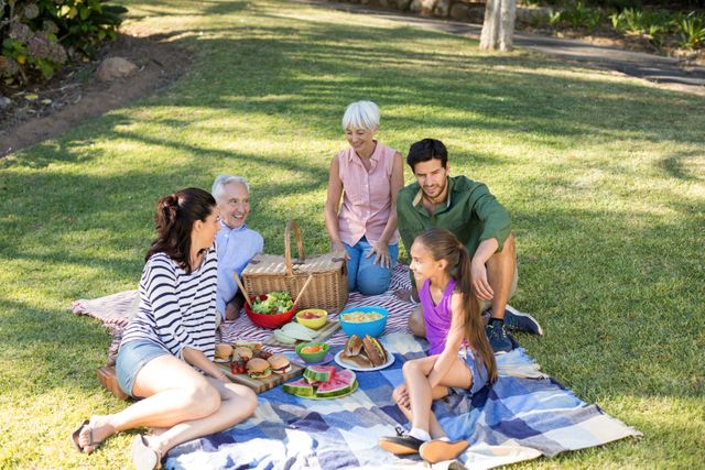Multi-generational family enjoying a picnic in a park on a sunny day. They are sitting on blankets with various foods laid out, including salads, sandwiches, and fruits. Good for illustrating family time, outdoor activities, healthy living, and bonding moments. Perfect for advertisements, blog posts, and articles about family gatherings, outdoor fun, and lifestyle.