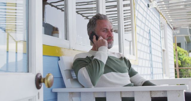 Mature man sits on porch, talking on smartphone. Suitable for themes related to leisure, communication, relaxation, tech use, and outdoors. Perfect for advertisements, lifestyle blogs, or digital communication topics.