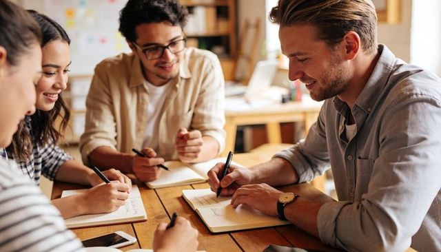 Young professionals collaborating and writing notes around sunlit wooden table in workspace