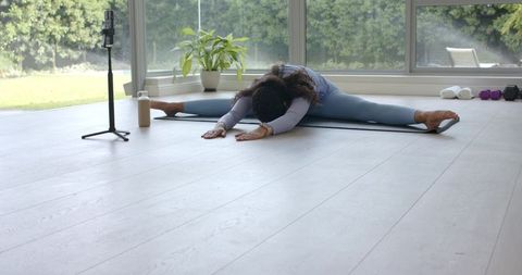 African american woman stretching straddle forward fold on yoga mat filming home workout