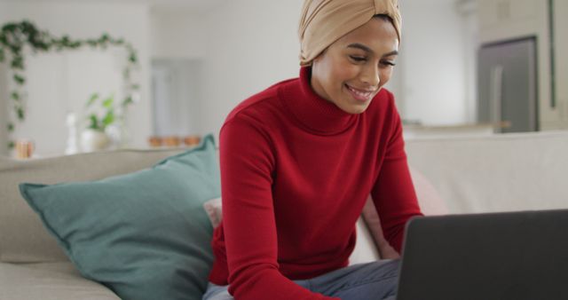 A woman wearing a hijab and red sweater is smiling while working on a laptop at home. The setting appears comfortable and modern with neutral tones and cozy cushions in the background. Ideal for representations of remote work, modern lifestyle, productive and comfortable home environment, technology use, or casual working atmospheres.