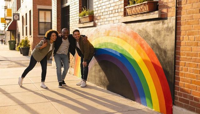Posing friends linking arms on sunlit urban sidewalk smiling by vibrant rainbow mural
