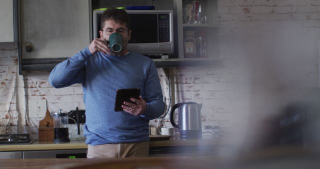Man standing in quaint kitchen sipping coffee and using tablet. Rustic ambiance features exposed brick and modern appliances. Ideal for illustrating morning routines, technology use at home, or cozy kitchen settings.
