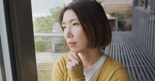 A woman with short hair, wearing a mustard cardigan, stands by a window in a modern house, looking outside thoughtfully. This can be used in mental health articles, blogs about introspection, personal growth, or mood-related topics. Useful for advertising lifestyle content or illustrating stories dealing with contemplation and self-reflection.