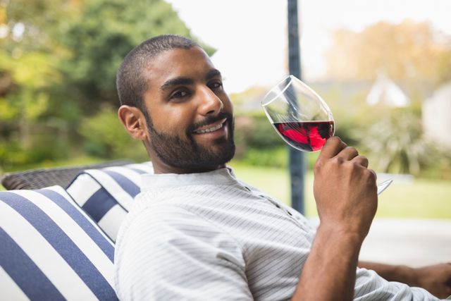 Man smiling while drinking red wine on porch, perfect for lifestyle blogs, advertisements for wine brands, leisure and relaxation themes, or promotional materials for outdoor furniture.
