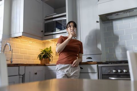 Asian woman leaning on kitchen counter drinking water in cozy modern home interior