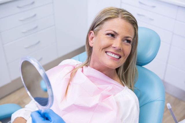 Smiling woman holding a mirror while sitting in a dental chair at a clinic. Ideal for use in healthcare, dental care, and patient satisfaction promotions. Can be used in advertisements for dental services, brochures, websites, and educational materials about oral health and hygiene.