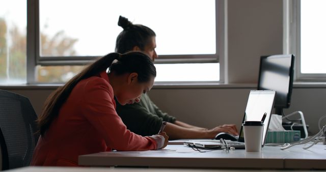 Two team members engaged in work at their desks in modern office environment. Great for illustrating professional office atmospheres, teamwork, collaborative workspaces, corporate settings, productivity, and modern workplace layouts.