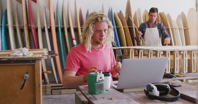 Two artisans shaping surfboards in workshop. One uses a laptop on wooden table while the other works in background. Surfboards and woodworking tools visible. Perfect for articles on craftsmanship, teamwork, and surfing culture.