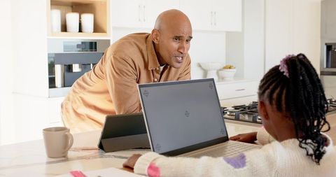 Dad guiding african american daughter using laptop at kitchen island for remote learning