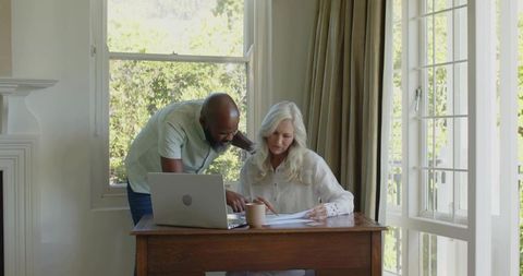 Couple reviewing finances and paperwork at home desk with laptop, daylight and garden view