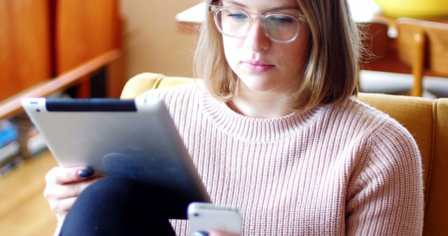 Woman wearing glasses and casual sweater using a tablet and smartphone indoors. Suitable for depicting modern technology use in everyday life, digital lifestyles, and multitasking. Ideal for articles and advertisements related to tech gadgets, remote work, or leisure time at home.