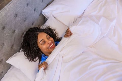 Smiling woman relaxing in bed holding white duvet wearing blue satin pajama top morning