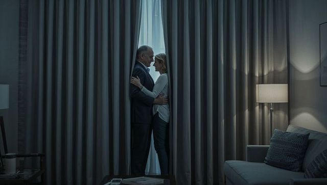 Mature couple sharing intimate embrace behind living room curtains with warm ambient lighting
