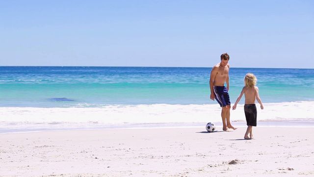 The scene shows a father and son enjoying a game of soccer on a sunlit beach with blue ocean waves in the background. This captures the essence of family bonding and outdoor activity, emphasizing leisure and playful moments. Suitable for concepts related to family life, vacations, sports, parent-child relationships, and cheerful outdoor settings. Ideal for use in advertising campaigns or content about family travel and sports activities.