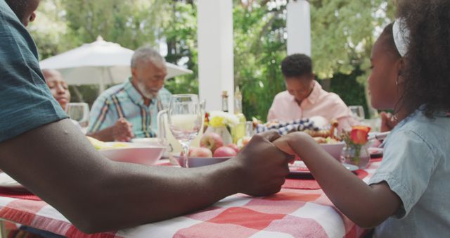 Happy african american family sitting at table in garden, praying together. Lifestyle, domestic life, family, and togetherness.