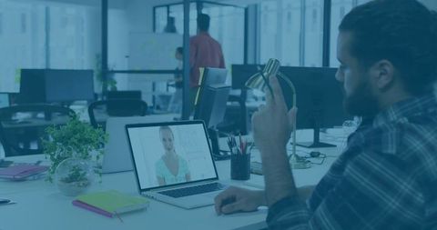 Bearded professional joining video call at modern open office desk with laptop and headset