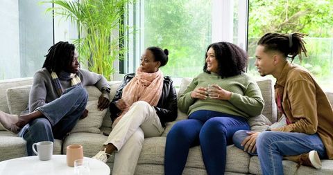 Diverse friends sitting and chatting on sunlit sofa with indoor greenery and coffee cups