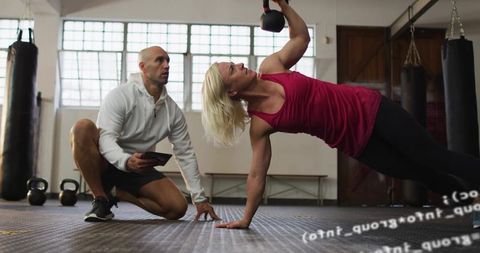 Female athlete performing kettlebell side plank while trainer monitoring progress with tablet