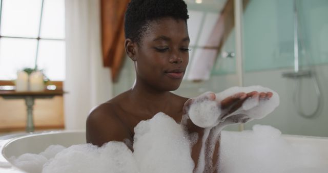 Black child enjoying bath time, playing with soap bubbles in modern bathroom. Perfect for content about childhood, hygiene habits, relaxation, self-care routines, and family lifestyle. Ideal for educational materials or wellness products marketing.