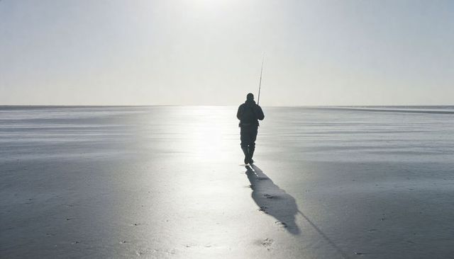 Solitary angler walking across backlit frozen shoreline with long shadow and footprints