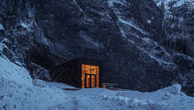 Glowing modern mountain shelter nestled under cliff, warm amber light in snowy night